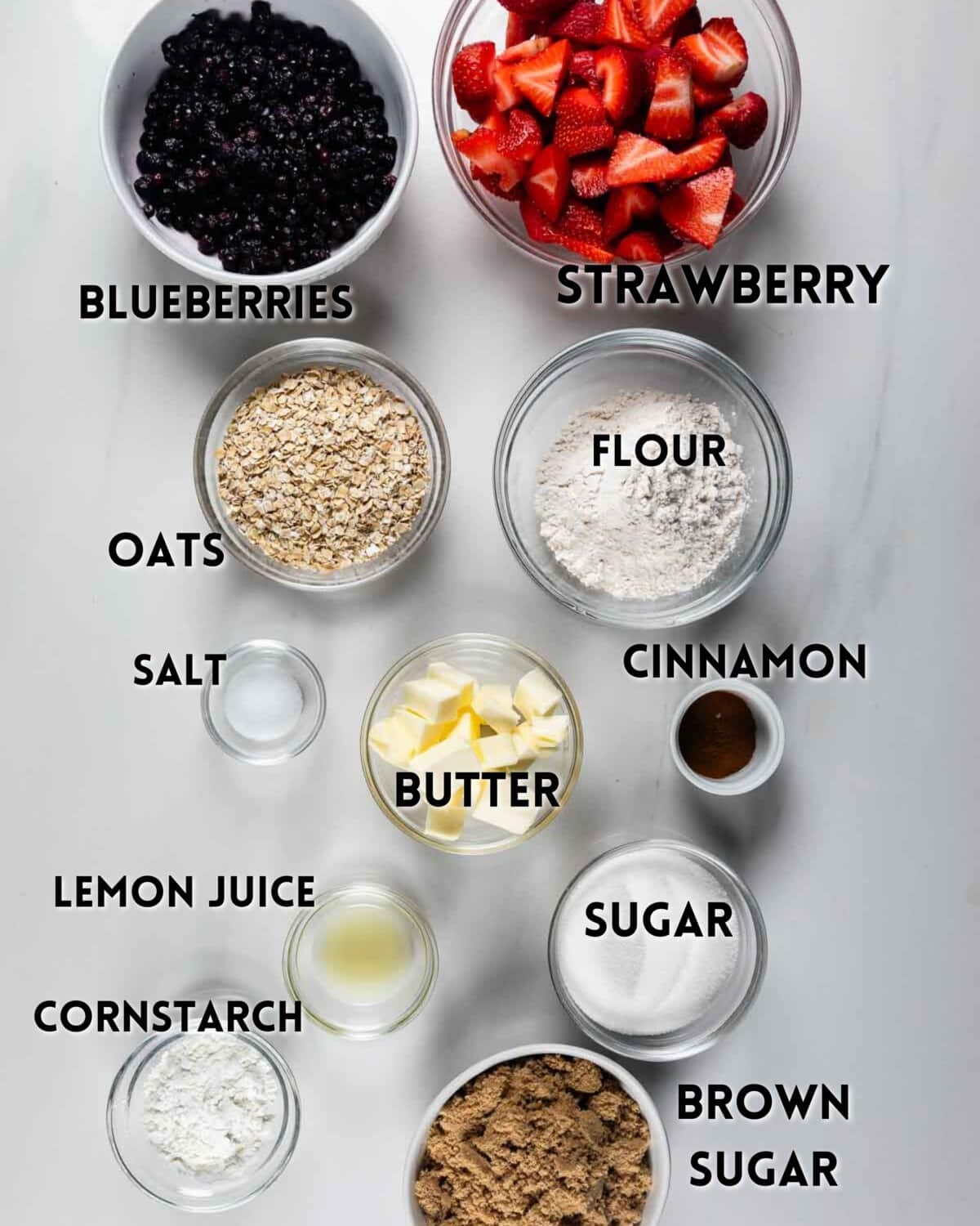 Various labeled ingredients in bowls on a white surface, including blueberries, strawberries, flour, oats, salt, cinnamon, butter, sugar, brown sugar, lemon juice, and cornstarch.