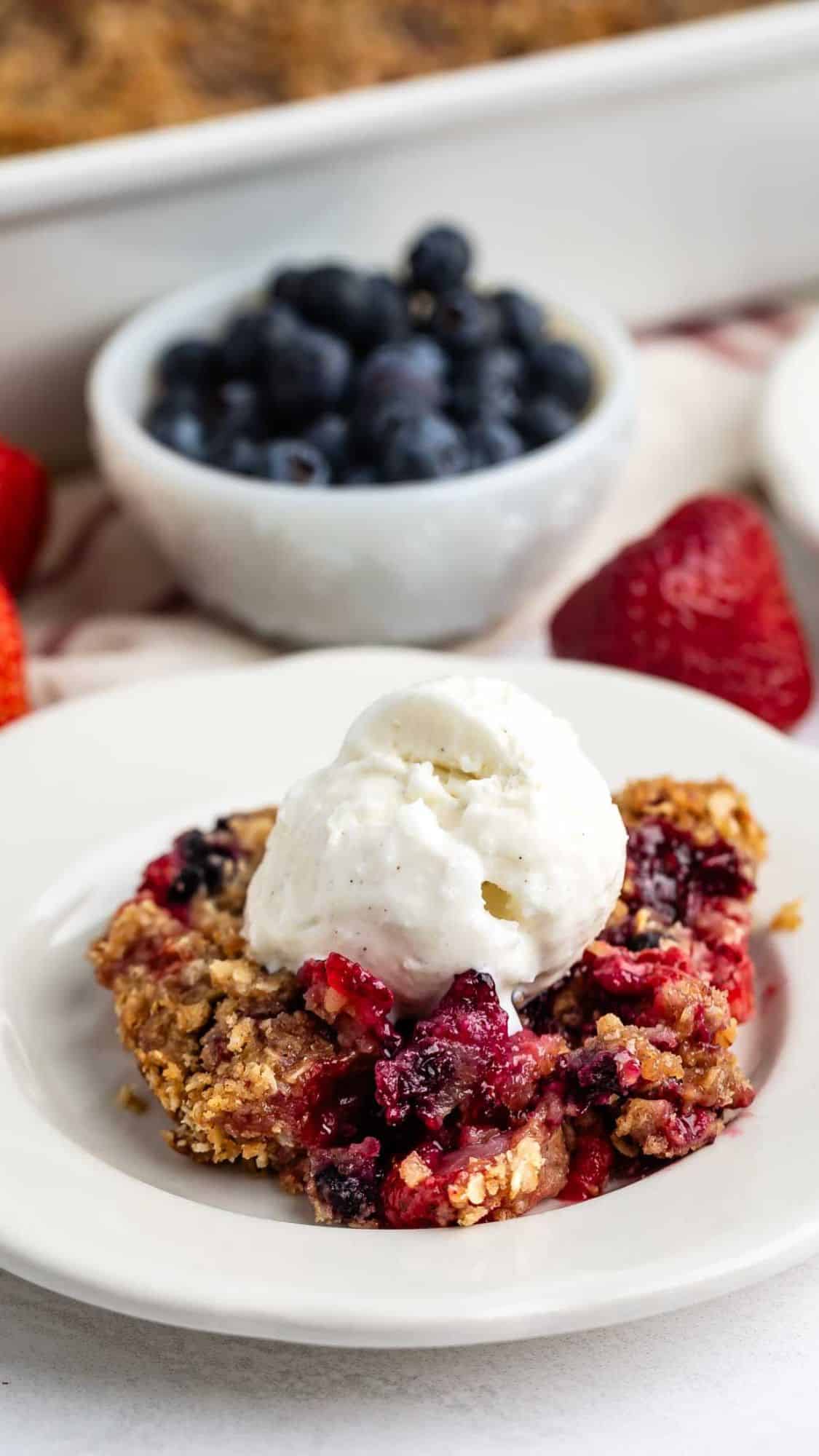A serving of mixed berry crumble topped with a scoop of vanilla ice cream on a white plate, with fresh blueberries and strawberries in the background.