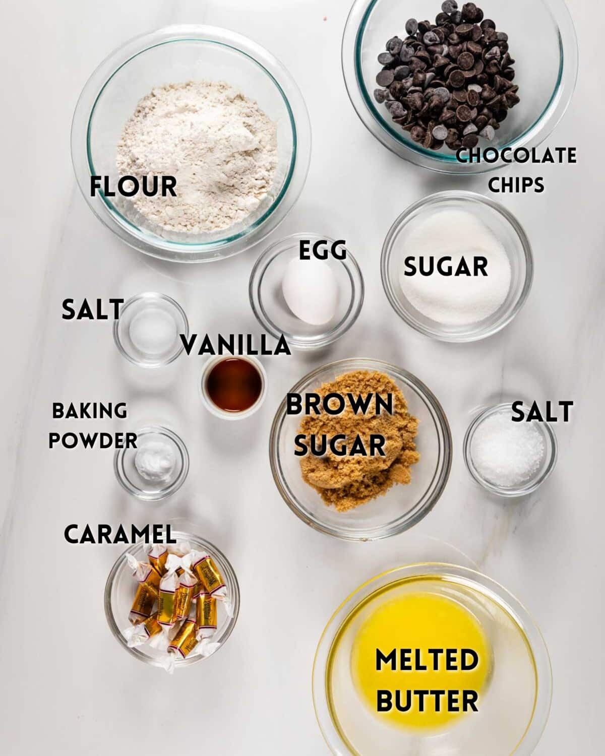 Several baking ingredients in glass bowls on a white surface, each labeled: flour, chocolate chips, egg, sugar, brown sugar, salt (appears twice), vanilla, baking powder, caramel candies, and melted butter.