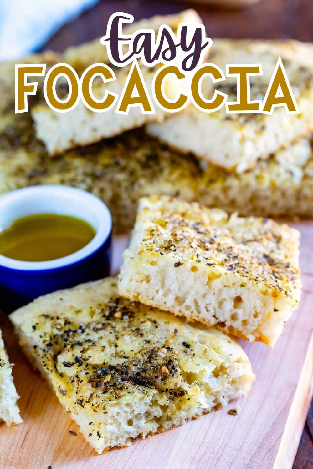 Close-up of thick, golden slices of focaccia bread topped with herbs, arranged on a wooden board with a small bowl of olive oil. Text above reads Easy Focaccia.