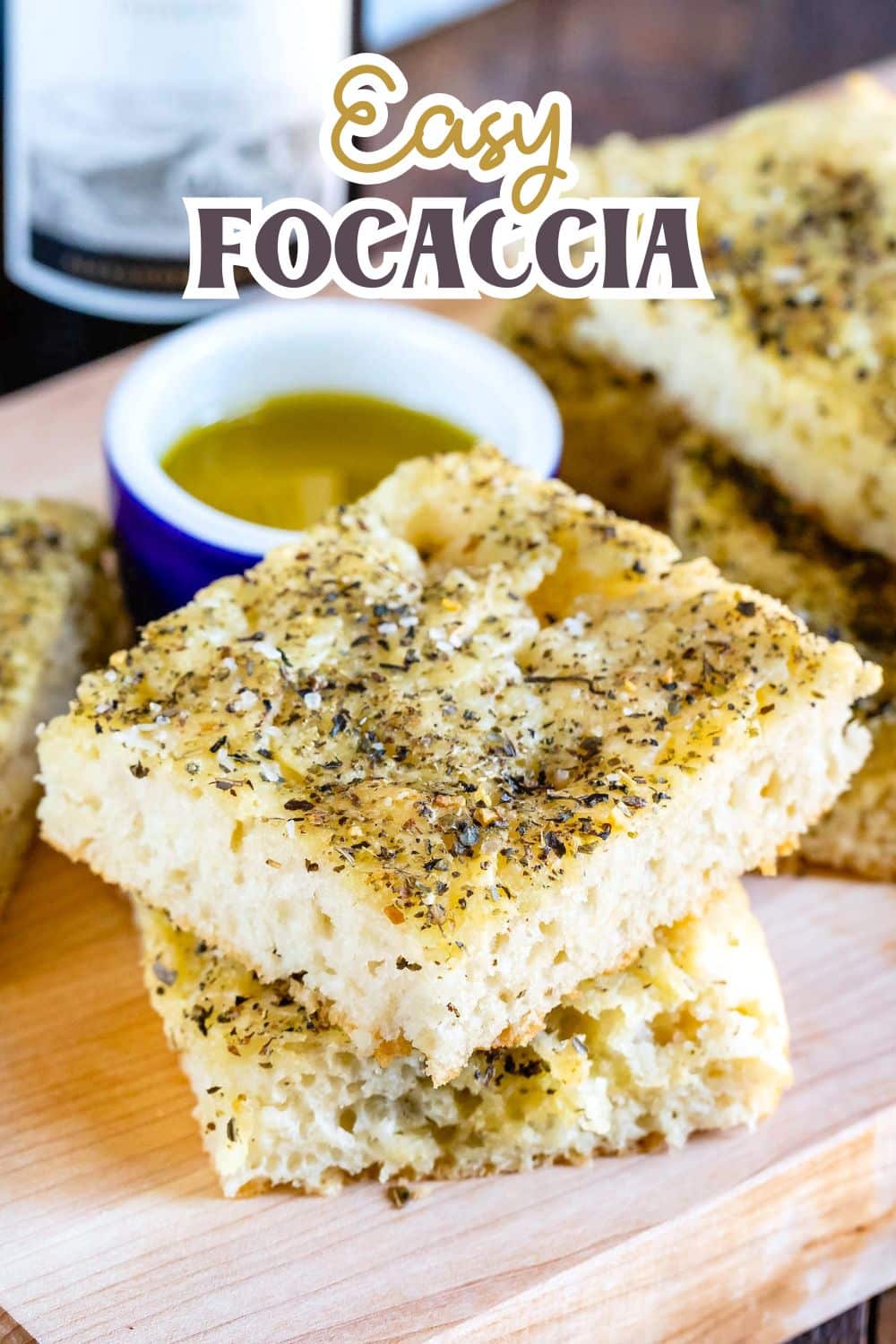 Sliced pieces of herb-topped focaccia bread stacked on a wooden board, with a small bowl of olive oil for dipping in the background. The text Easy Focaccia appears at the top of the image.