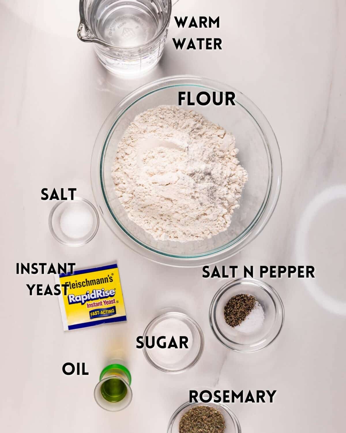 A bowl of flour at the center, surrounded by labeled ingredients: warm water, salt, instant yeast, oil, sugar, salt and pepper, and rosemary, all arranged on a white surface.