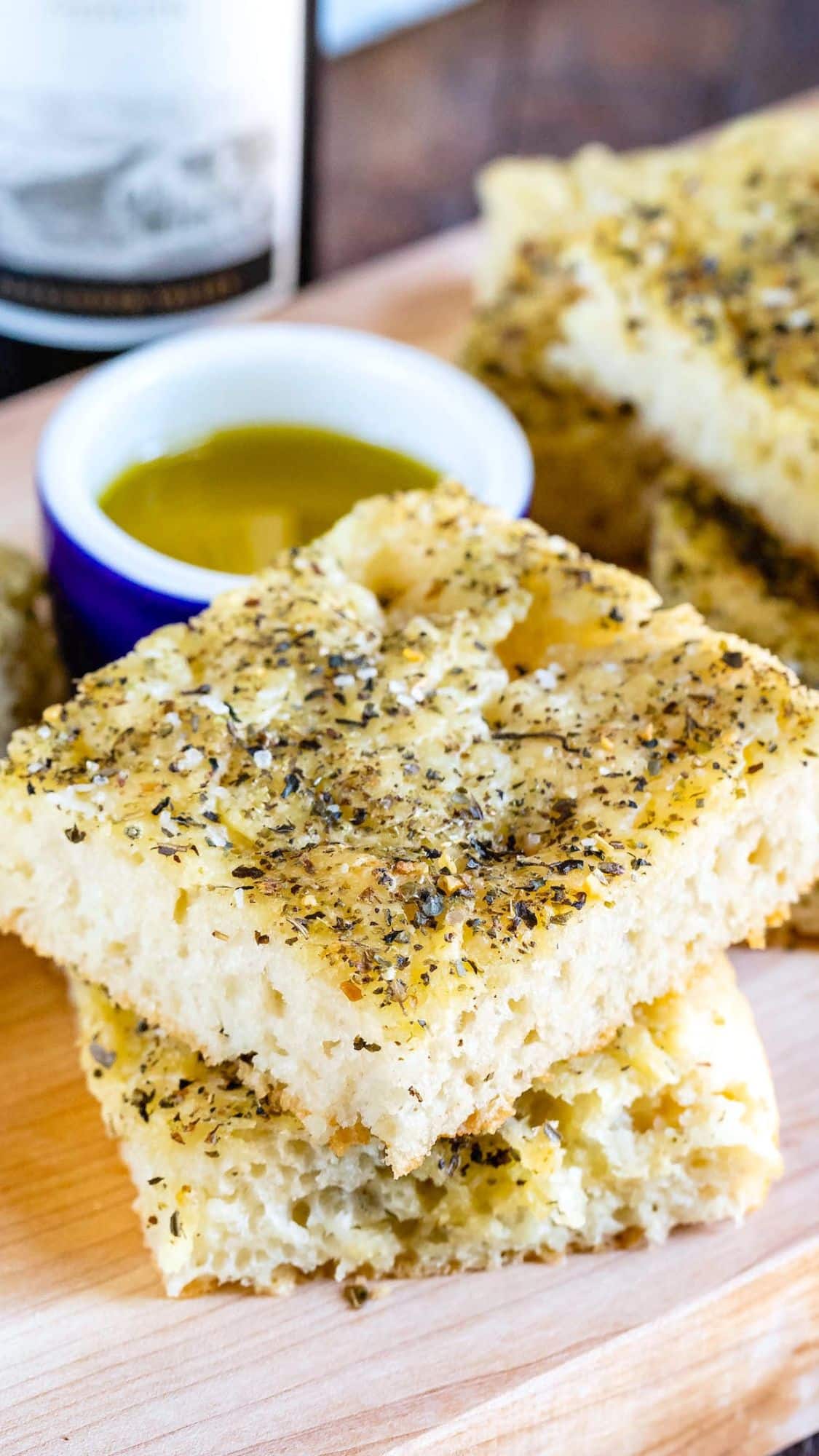 Two pieces of herbed focaccia bread stacked on a wooden board, with a small dish of olive oil for dipping in the background. The bread is topped with dried herbs and looks fluffy and golden.