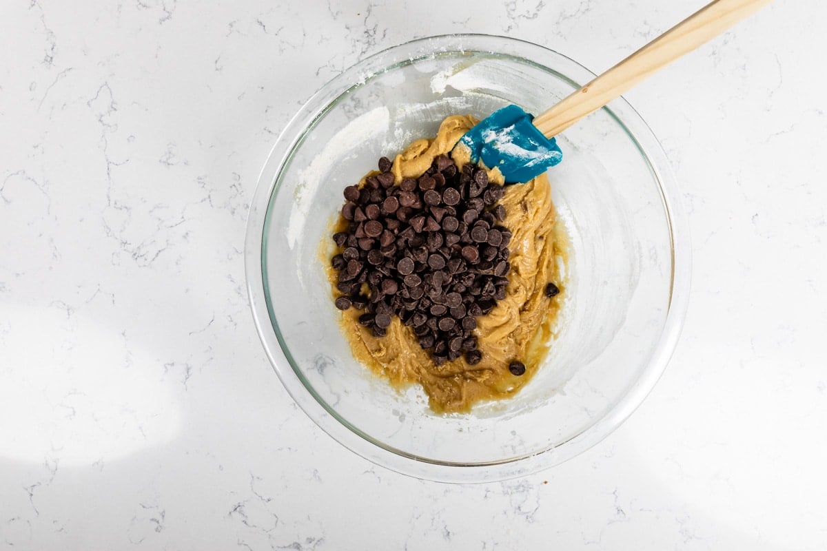 A glass bowl containing cookie dough with chocolate chips and a blue spatula rests on a white marble countertop.