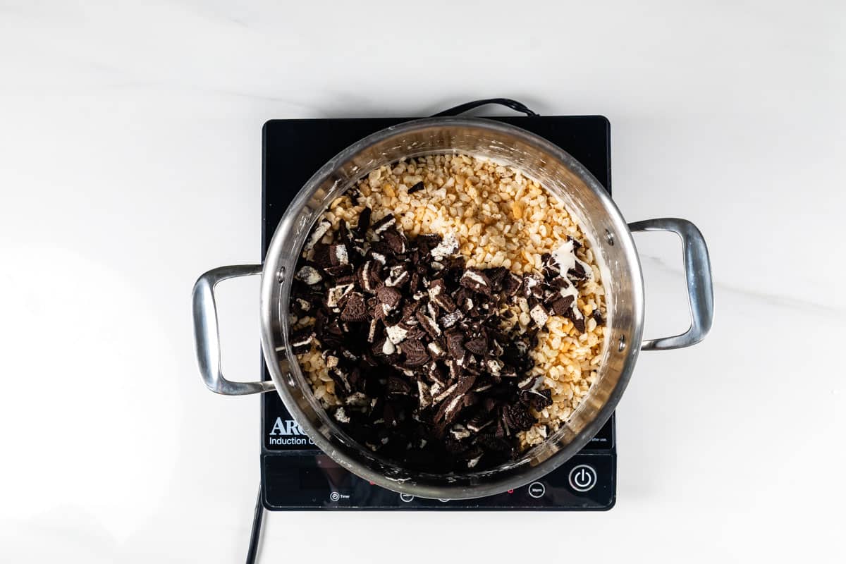 A pot on an induction cooktop contains Rice Krispies cereal and crushed chocolate sandwich cookies, viewed from above, ready to be mixed together to make a dessert.