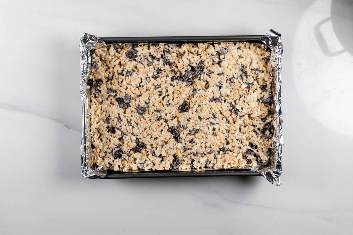 A rectangular baking pan lined with aluminum foil is filled with an unbaked cookie or dessert bar mixture, containing visible chunks of dark chocolate or dried fruit. The pan sits on a light-colored surface.