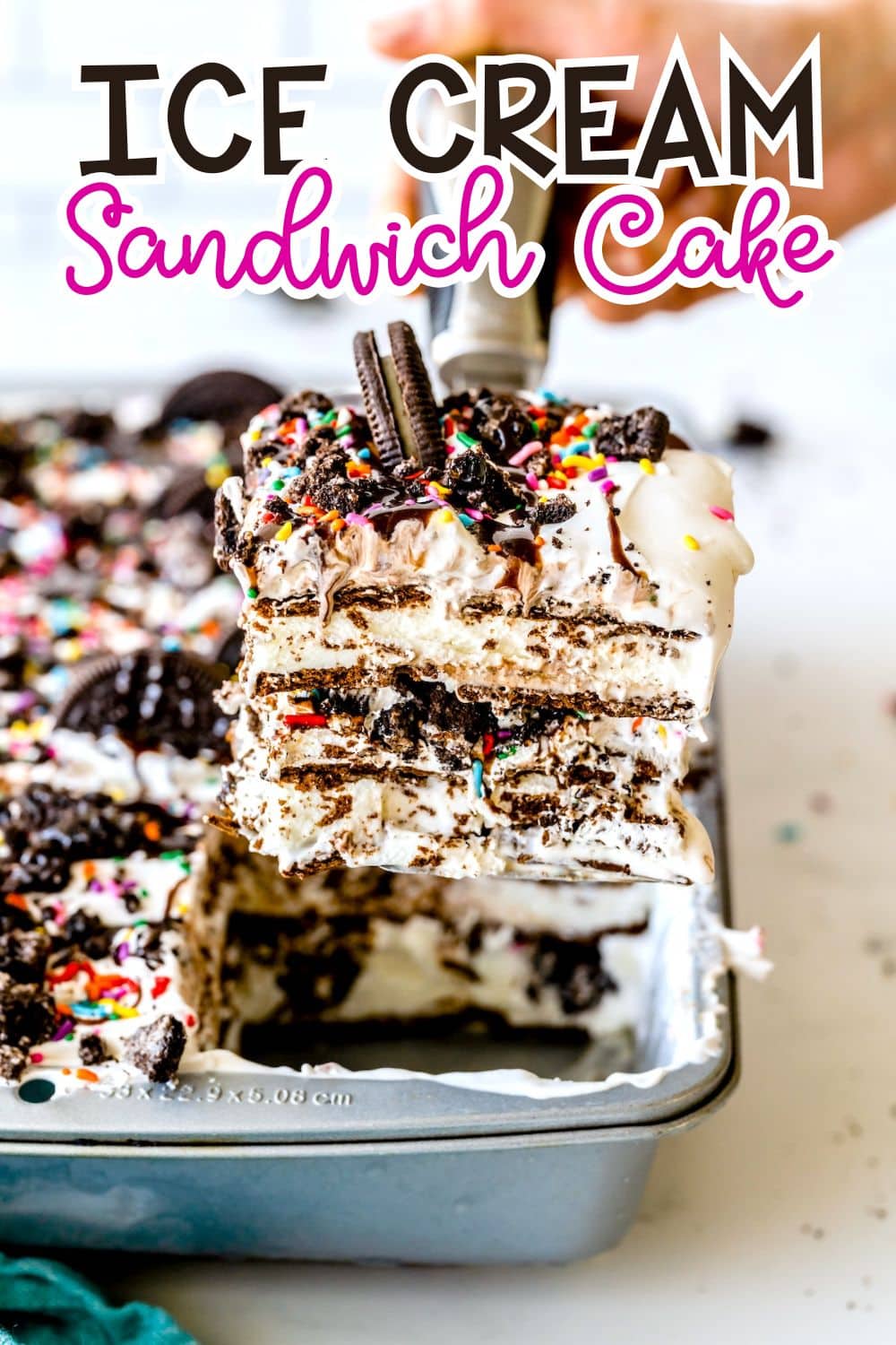 A spatula lifts a large square piece of ice cream sandwich cake topped with whipped cream, sprinkles, and crushed cookies from a baking pan, with the rest of the cake still inside the pan.