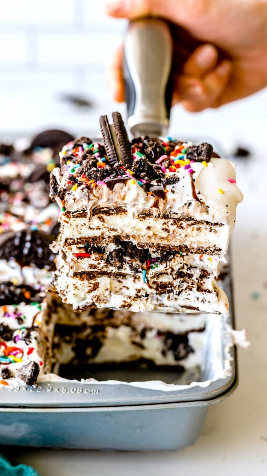 A hand lifts a layered ice cream cake with cookies, whipped cream, chocolate drizzle, and colorful sprinkles from a pan. Whole and crumbled cookies decorate the top.
