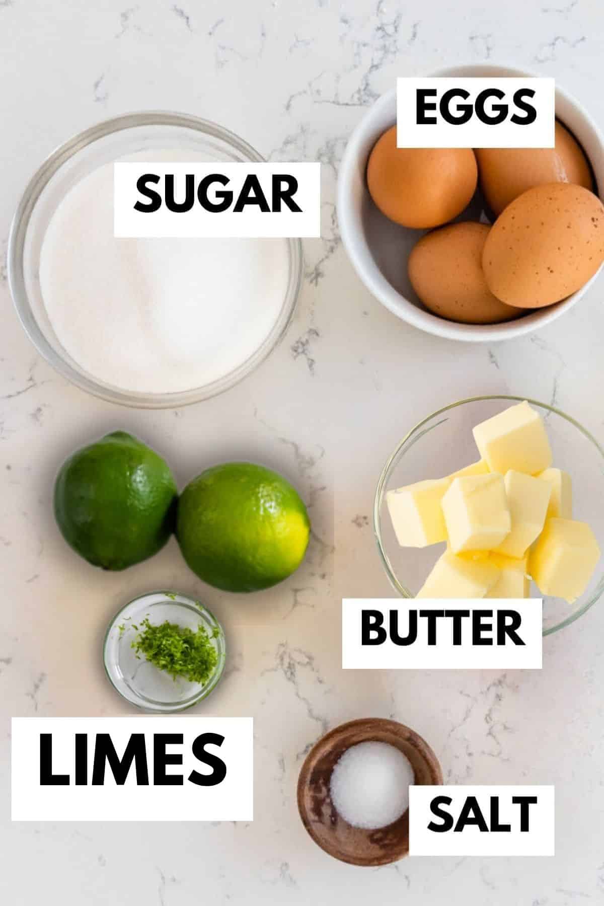 Top-down view of ingredients on a white surface, labeled: a bowl of sugar, a bowl of eggs, two limes, a bowl of butter cubes, a small bowl of lime zest, and a small bowl of salt.