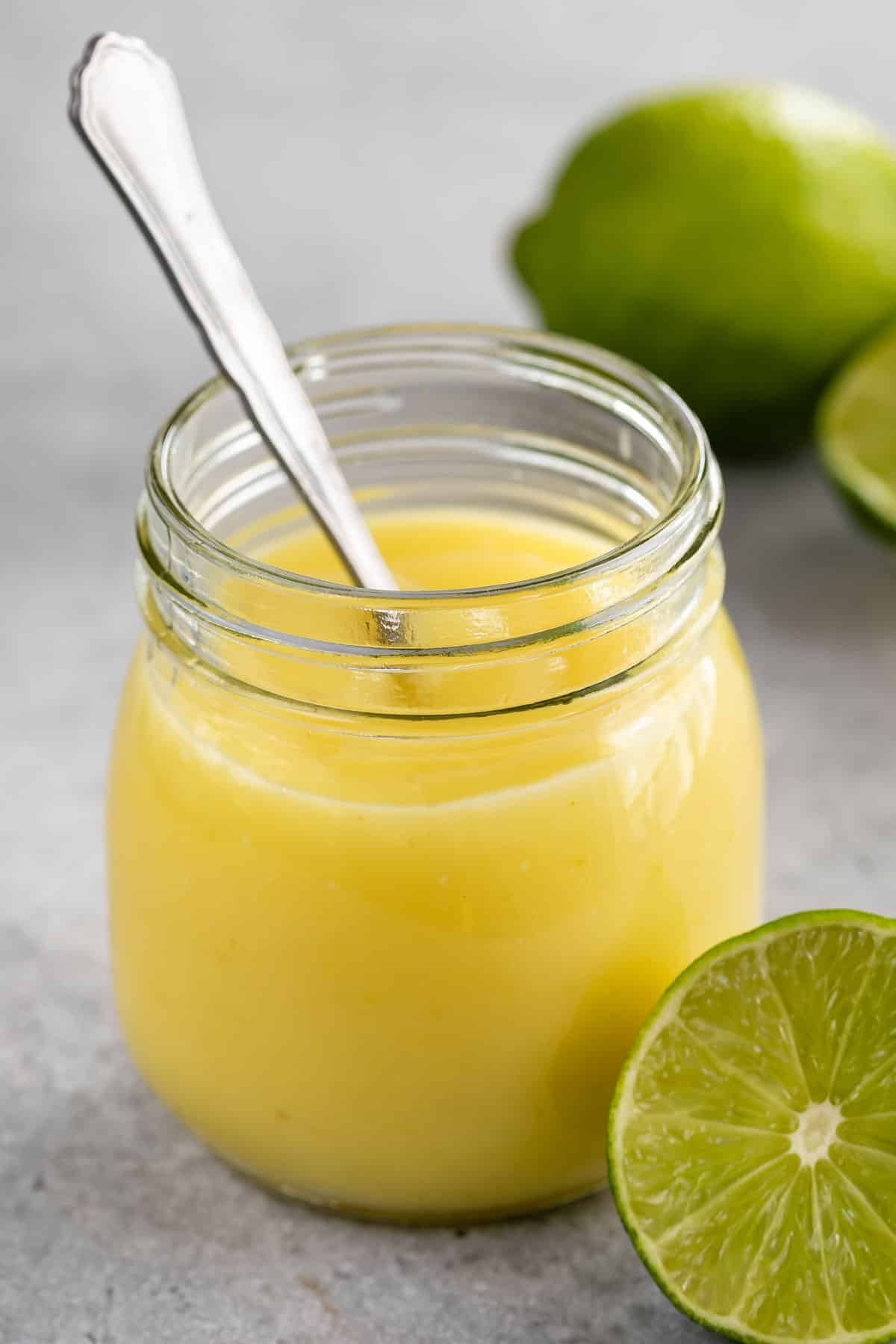 A glass jar filled with creamy yellow lime curd, with a metal spoon inside. In the background, there are whole and halved limes on a light gray surface.