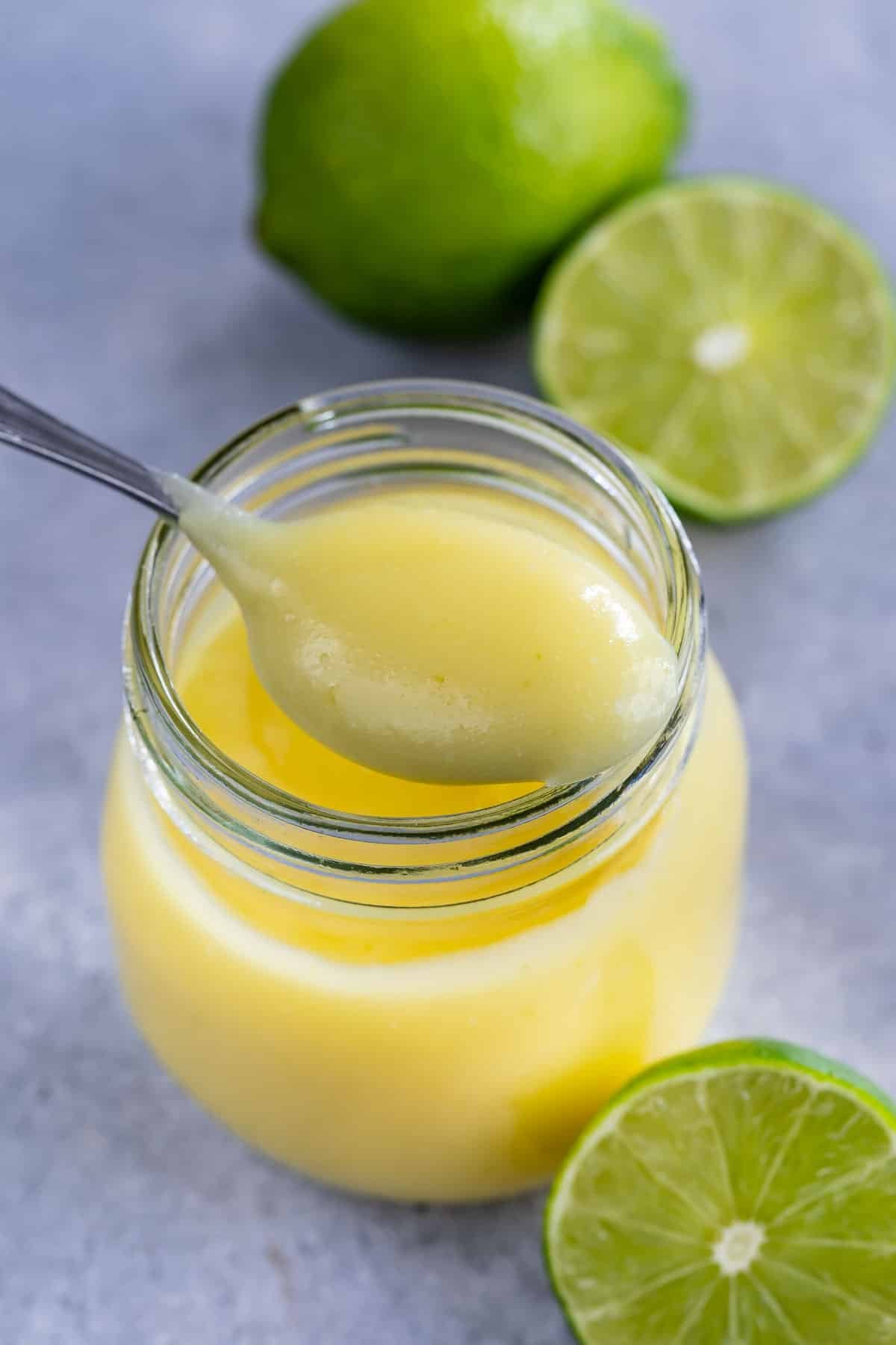A spoonful of creamy yellow lime curd is held above a glass jar, with fresh whole and halved limes in the background on a gray surface.