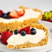 cheesecake topped with frosting, blueberries, and strawberries with the fruits also sprinkled around the cheesecake.