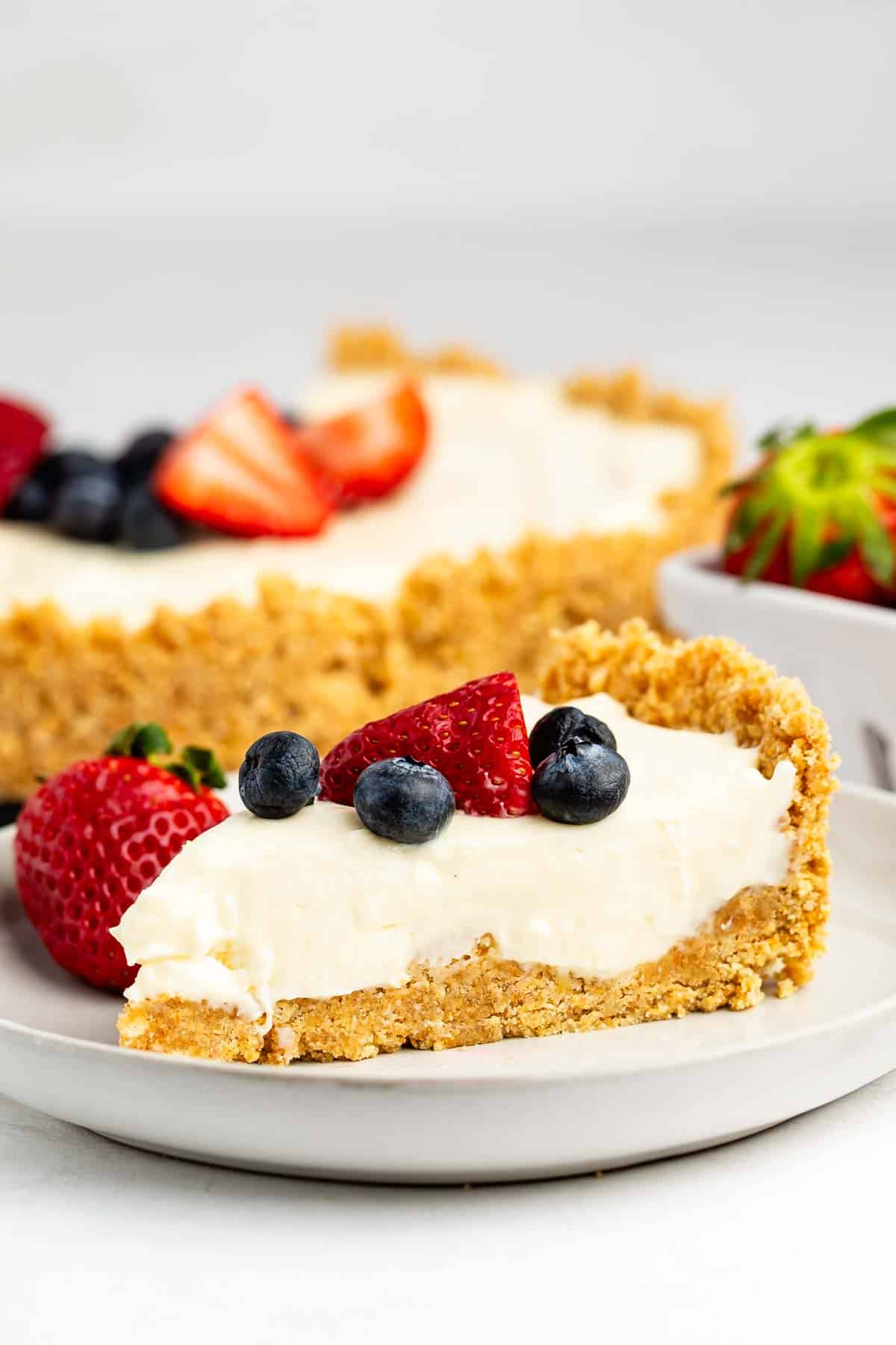 cheesecake topped with frosting, blueberries, and strawberries with the fruits also sprinkled around the cheesecake.