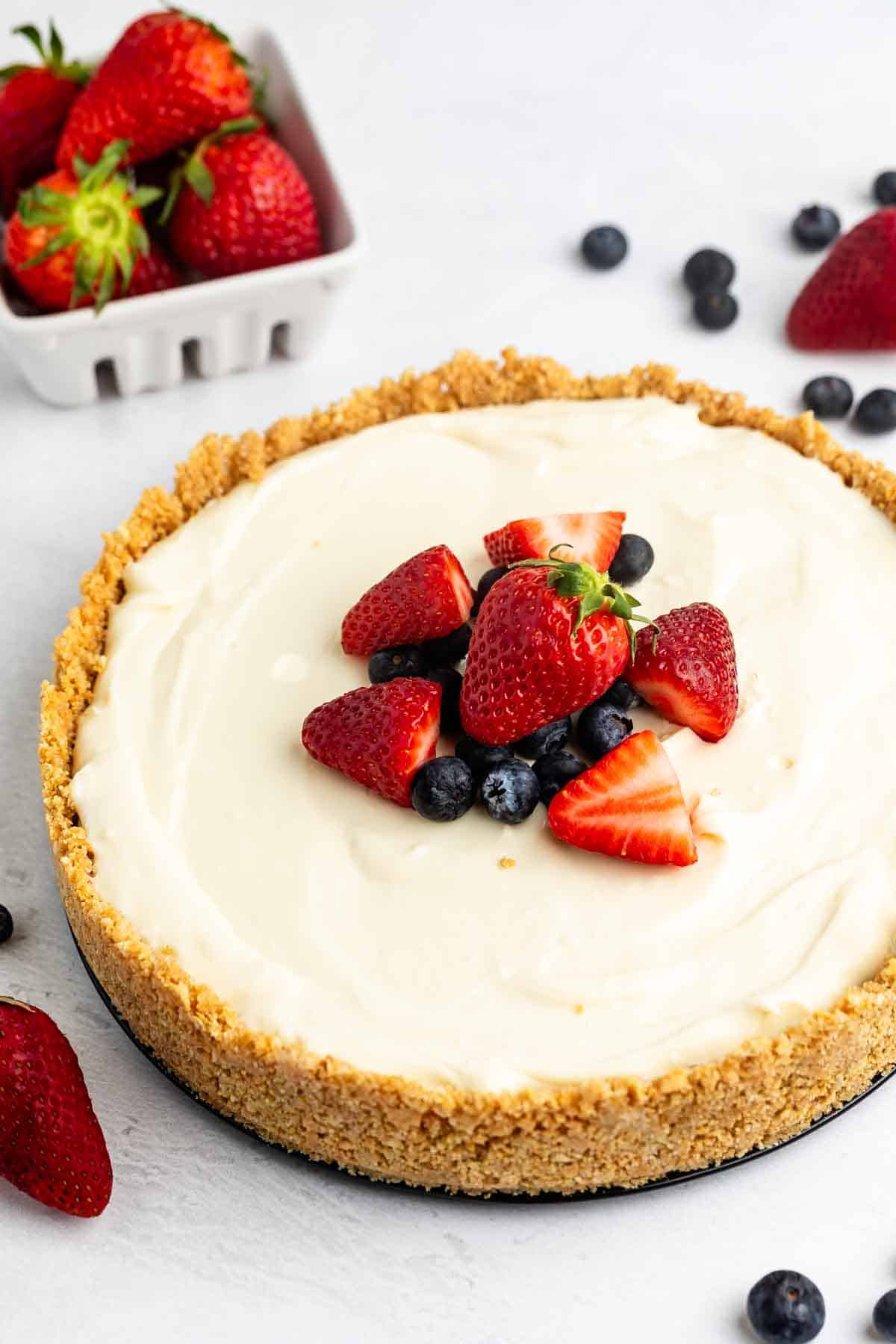 cheesecake topped with frosting, blueberries, and strawberries with the fruits also sprinkled around the cheesecake.