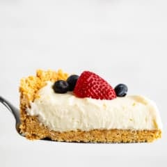 cheesecake topped with frosting, blueberries, and strawberries with the fruits also sprinkled around the cheesecake.