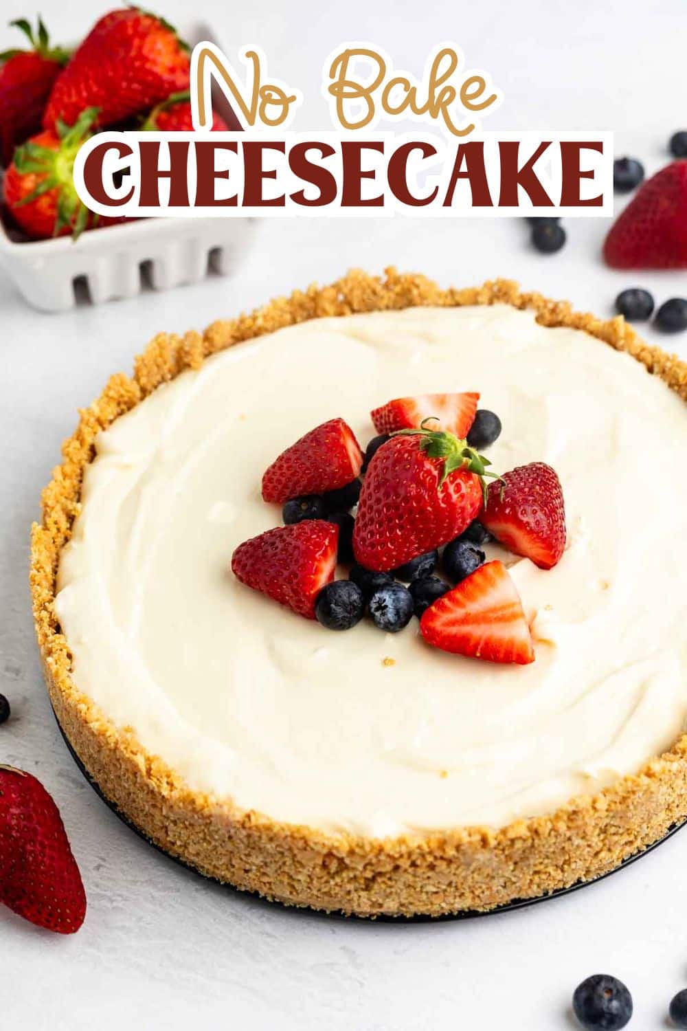 cheesecake topped with frosting, blueberries, and strawberries with the fruits also sprinkled around the cheesecake with words on the image that read the name of the recipe.