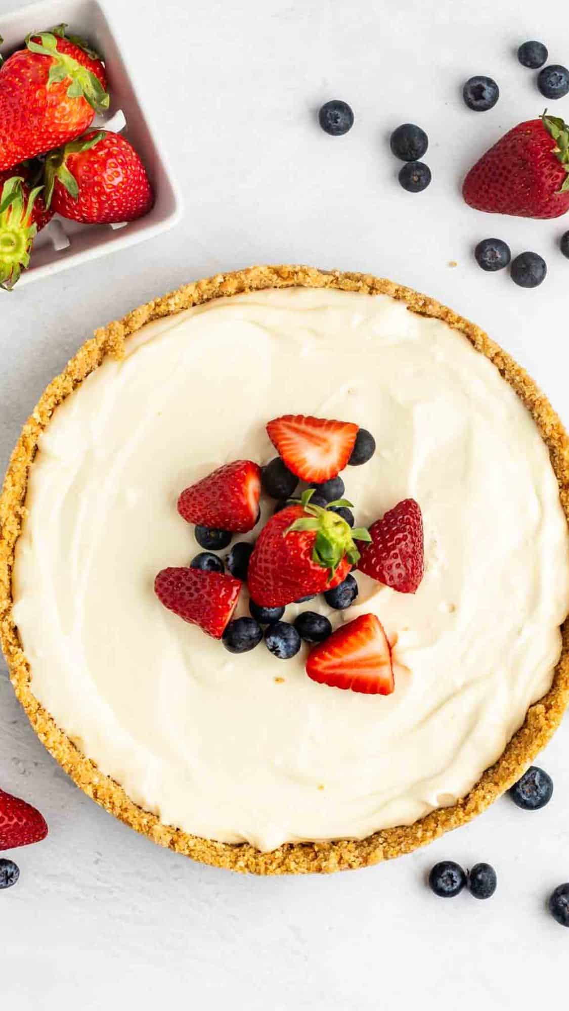 cheesecake topped with frosting, blueberries, and strawberries with the fruits also sprinkled around the cheesecake.