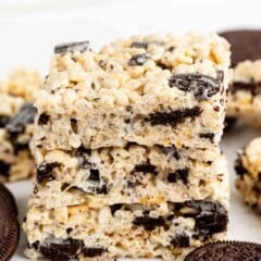 Three stacked rice krispie treats with visible chunks of chocolate sandwich cookies, surrounded by whole cookies on a white surface.