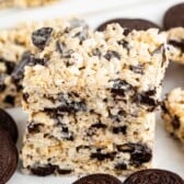 A close-up of three stacked rice crispy treats mixed with crushed chocolate sandwich cookies, with whole cookies scattered around on a white surface.