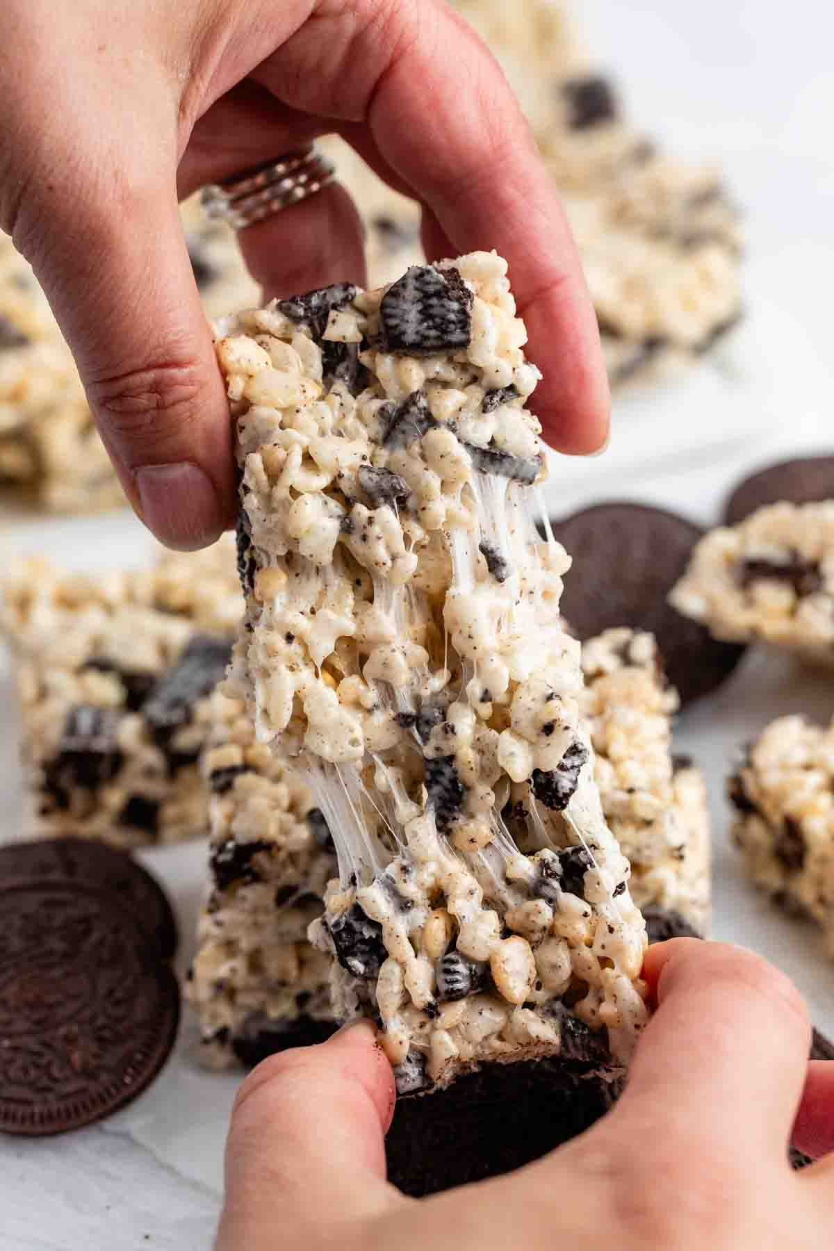 A close-up of hands pulling apart a gooey cookies and cream rice crispy treat, with visible chunks of chocolate cookies and marshmallow strands stretching between the pieces. More treats and sandwich cookies are blurred in the background.