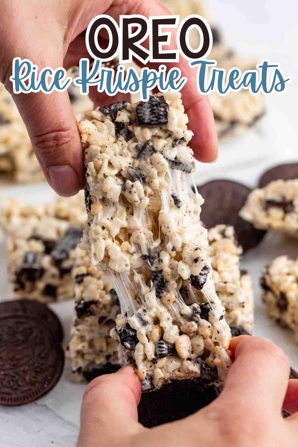 Close-up of hands pulling apart a gooey Oreo Rice Krispie Treat, showing melted marshmallow strands. Other treats and Oreo cookies are visible in the background. Text reads OREO Rice Krispie Treats at the top.