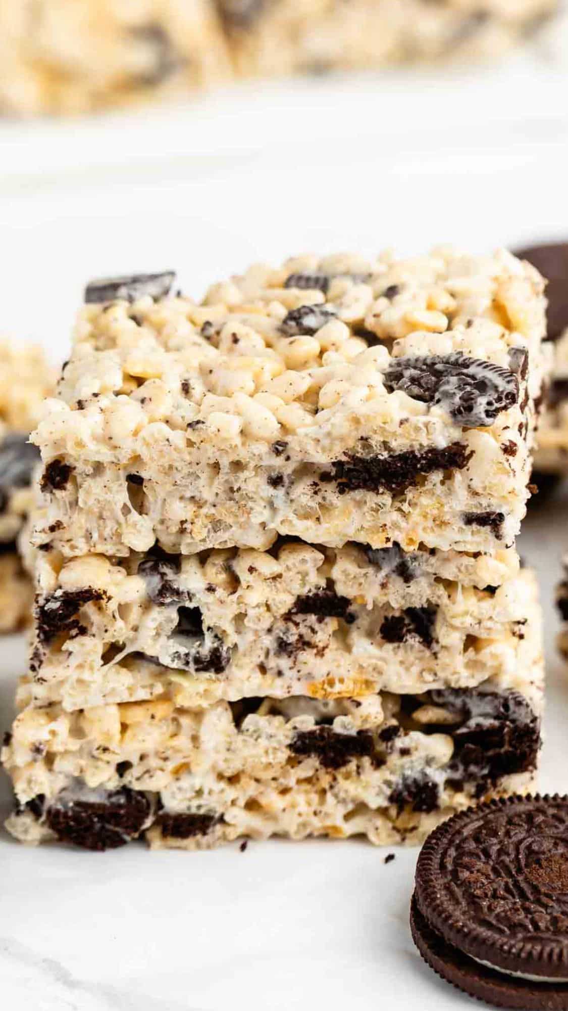 A close-up of three stacked rice krispie treats with crushed chocolate sandwich cookies mixed in, with whole cookies visible nearby on a white surface.