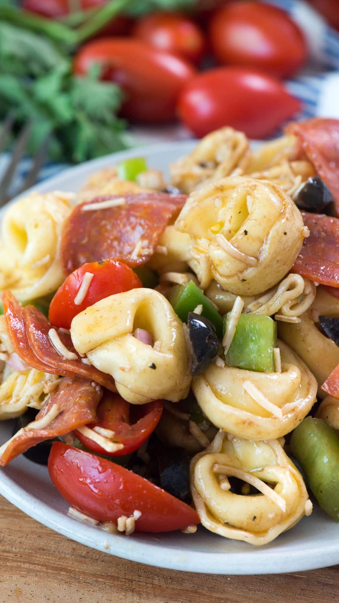 A close-up of a pasta salad with cheese tortellini, sliced pepperoni, cherry tomatoes, black olives, green bell peppers, and shredded cheese on a plate.