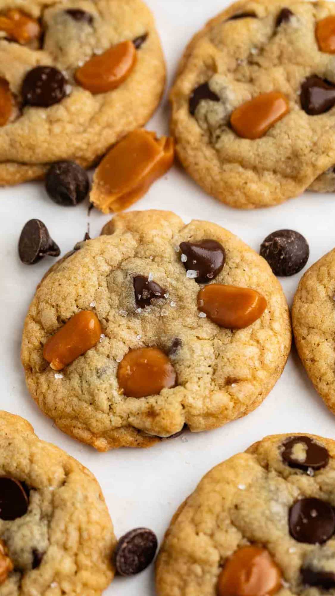 Close-up of freshly baked cookies with chocolate chips and caramel chunks, sprinkled lightly with sea salt, arranged on a white surface.