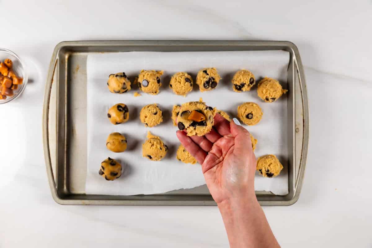 A hand holds a ball of chocolate chip cookie dough with caramel pieces inside, above a baking tray lined with parchment paper and rows of cookie dough balls. A bowl of caramels sits nearby.