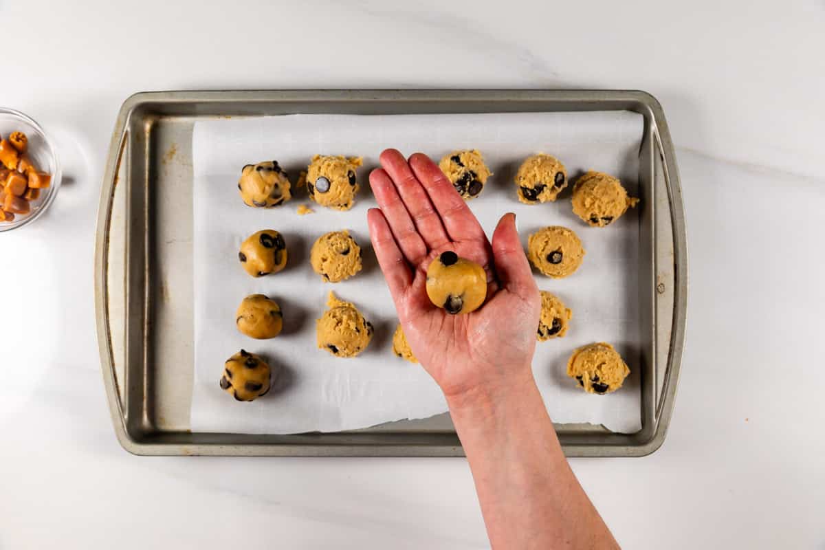 A hand holds a ball of chocolate chip cookie dough above a baking tray lined with parchment paper and rows of dough balls. A small bowl of caramel pieces sits nearby.