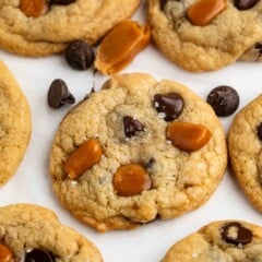 A close-up of several cookies studded with chocolate chips and caramel pieces, sprinkled with coarse sea salt, on a white surface. Loose chocolate chips and caramel bits are scattered around.
