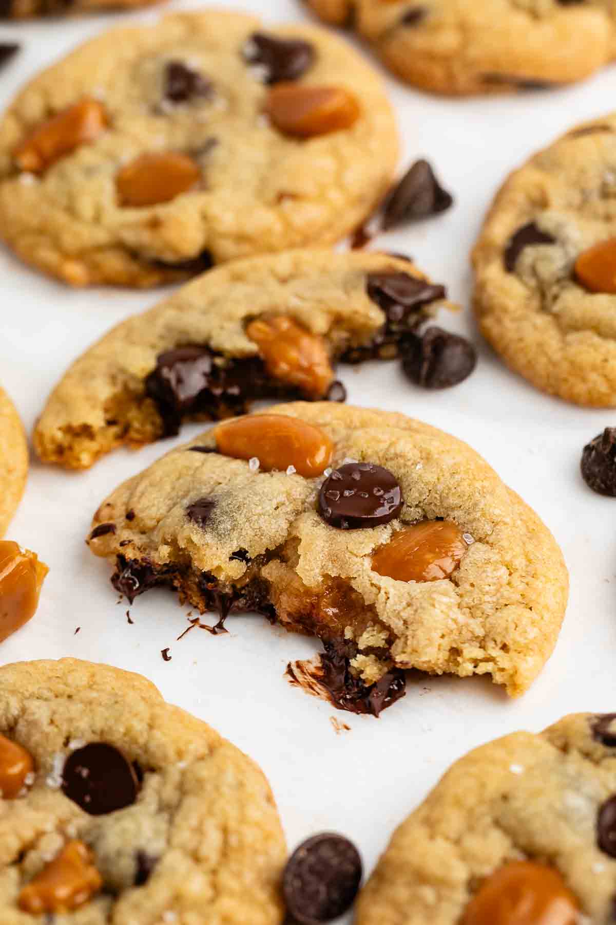 A close-up of chocolate chip cookies with caramel chunks, some with melted chocolate visible and one cookie partially eaten, all on a white surface.