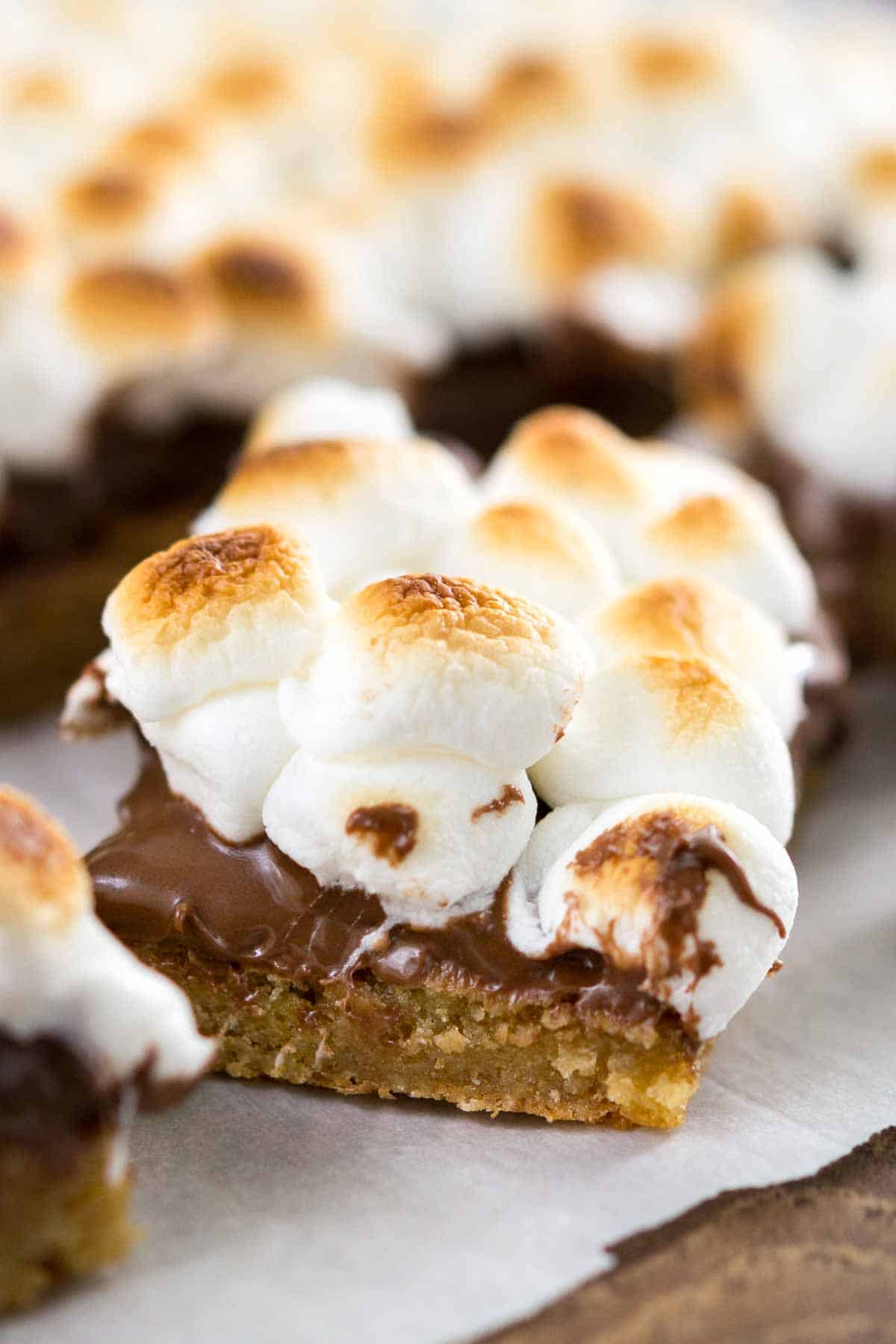 A close-up of a dessert bar with a golden cookie base, topped with a layer of melted chocolate and toasted mini marshmallows. Other similar dessert bars are visible in the background.