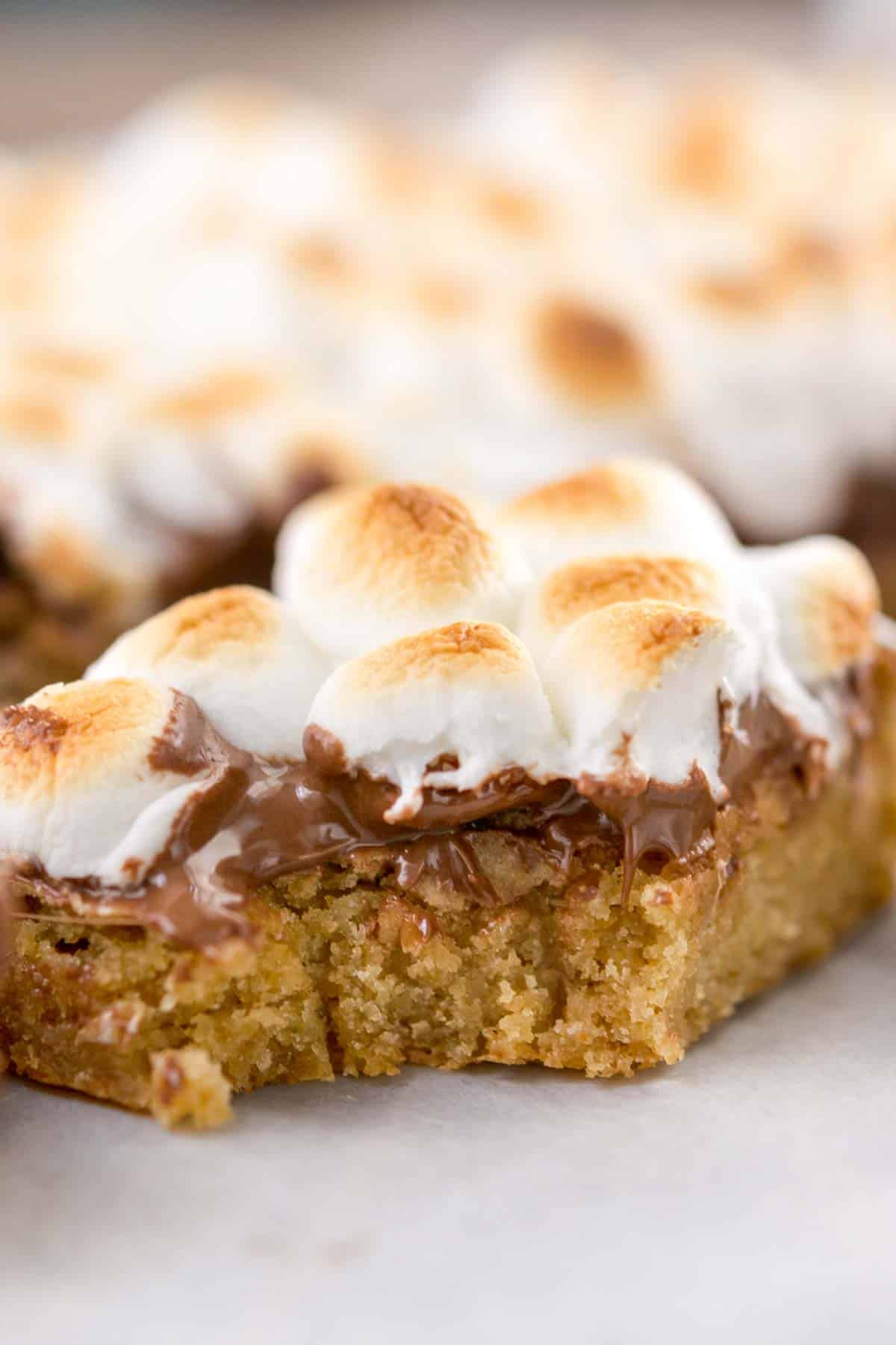 A close-up of a gooey dessert bar topped with toasted marshmallows and melted chocolate on a golden brown cookie base. The bar has a bite taken out of it, revealing its soft texture.