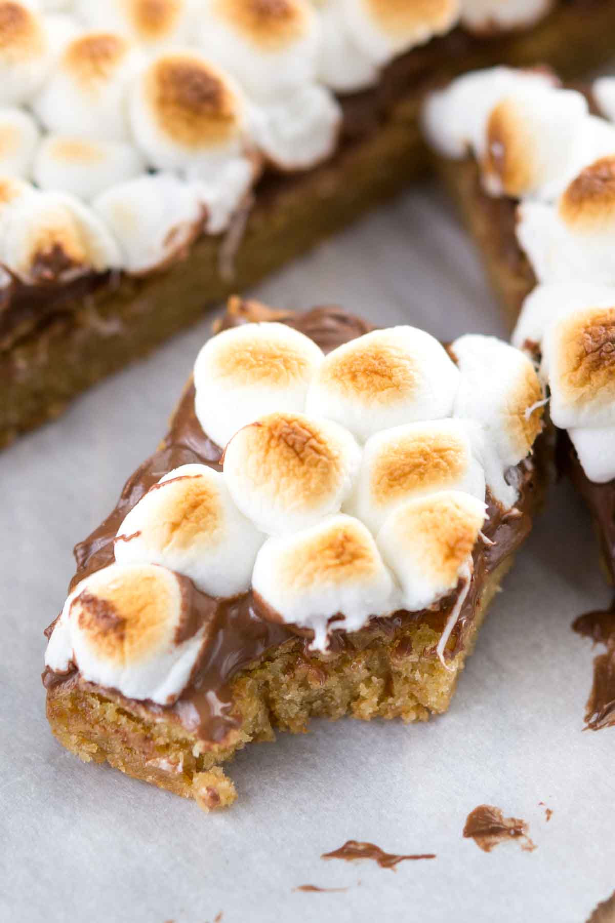 A close-up of a dessert bar topped with melted chocolate and toasted mini marshmallows, with a bite taken out of it. Other similar dessert bars are visible in the background on parchment paper.