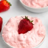 A white bowl filled with fluffy pink strawberry mousse, topped with a fresh whole strawberry. Another bowl of mousse and whole strawberries are visible in the background.