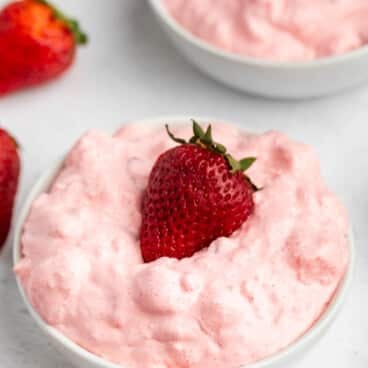 A white bowl filled with fluffy pink strawberry mousse, topped with a fresh whole strawberry. Another bowl of mousse and whole strawberries are visible in the background.