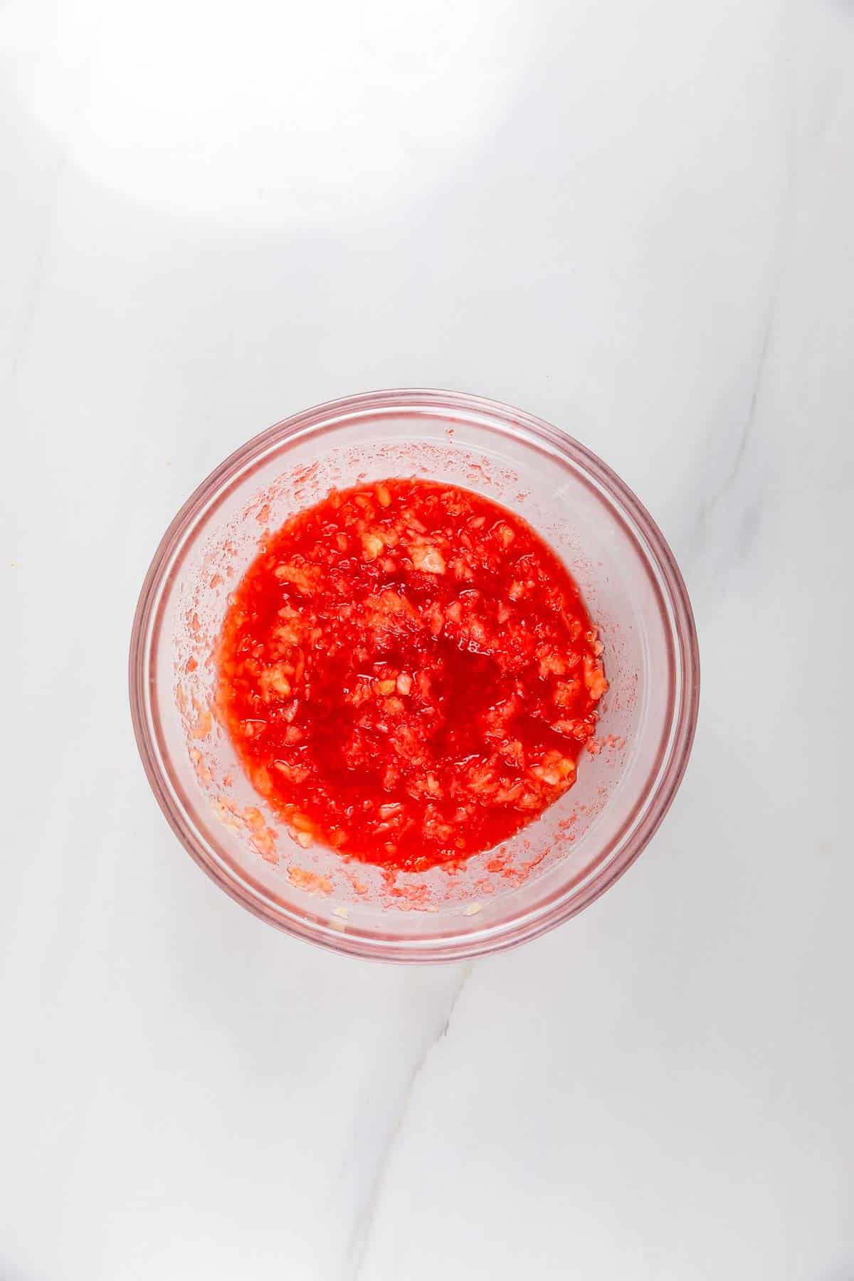 A clear glass bowl filled with a chunky red sauce or salsa, placed on a white marble surface.