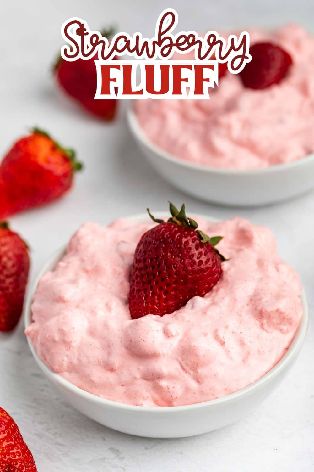 A white bowl filled with creamy pink strawberry fluff dessert, topped with a whole strawberry, sits on a white surface with more strawberries and another bowl of fluff in the background. The text Strawberry Fluff appears at the top.