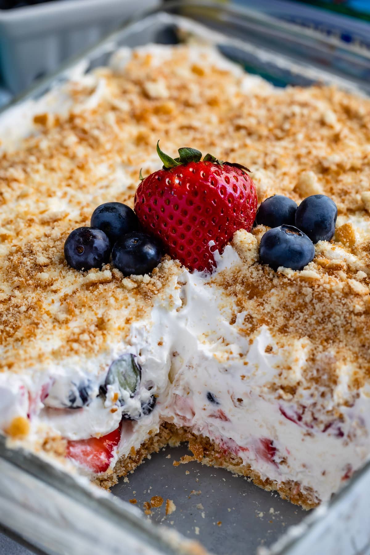 A creamy dessert in a glass dish topped with crumbled crumbs, fresh blueberries, and a strawberry. A piece is missing, revealing layers of whipped filling and fruit inside.