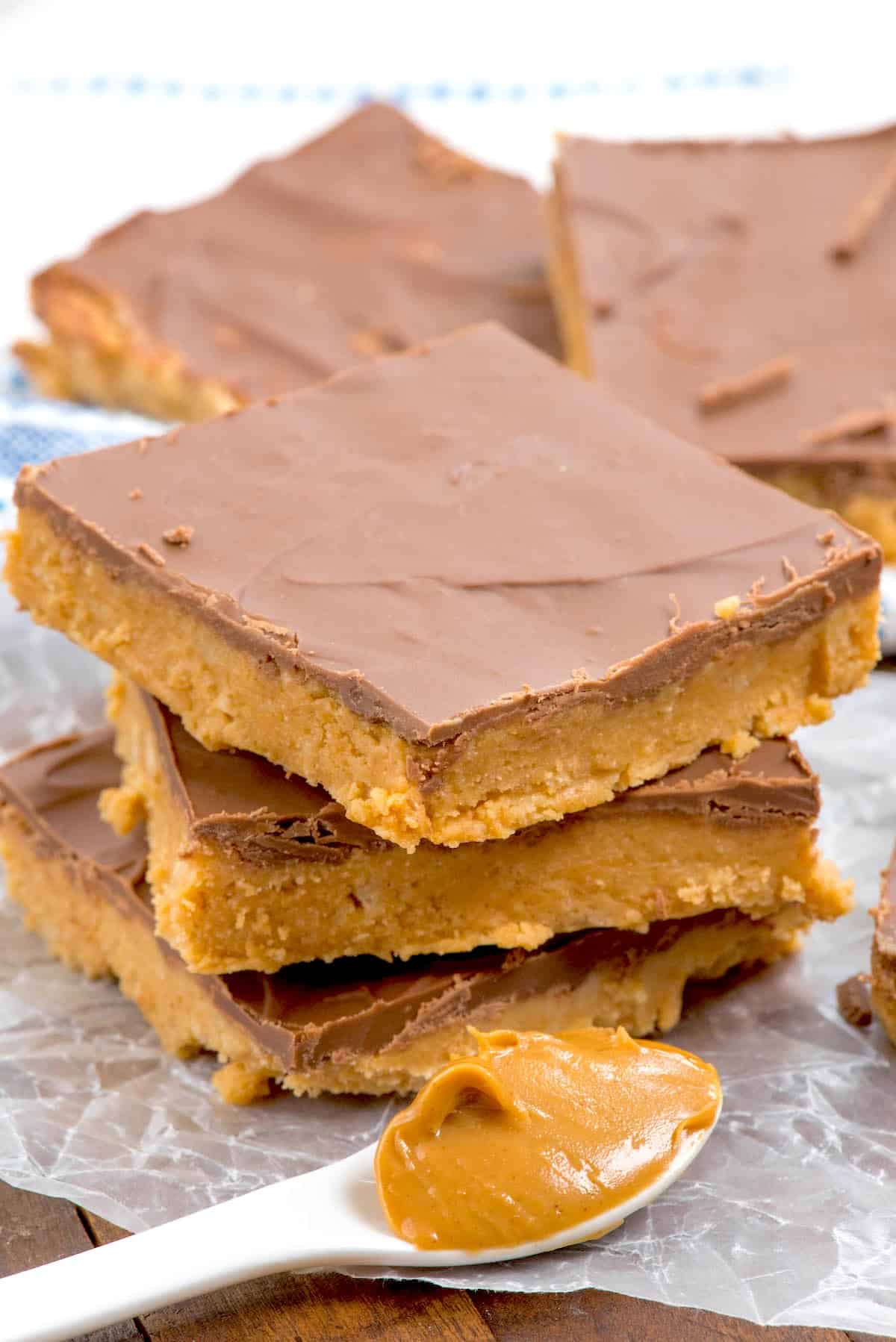 Three peanut butter bars with a chocolate topping are stacked on wax paper, with more bars in the background. A spoonful of peanut butter rests in the foreground.