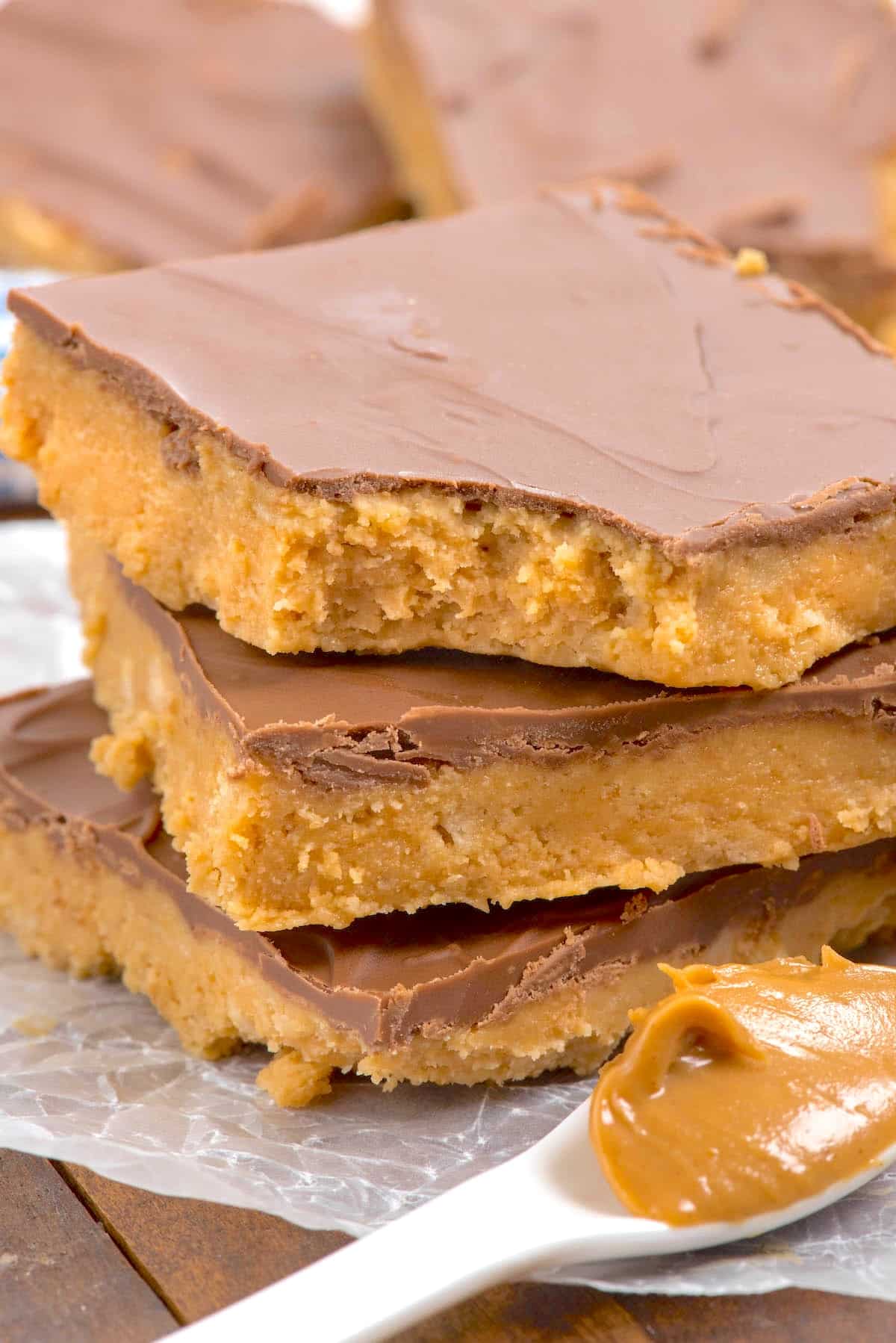 Three peanut butter bars with a chocolate topping are stacked on parchment paper. A spoonful of creamy peanut butter sits in the foreground. The bars have a thick, crumbly base and smooth chocolate layer.