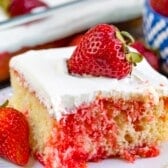 A slice of strawberry poke cake with white frosting and a whole strawberry on top, sitting on a white plate. The cake has visible red strawberry gelatin throughout and fresh strawberries beside it.