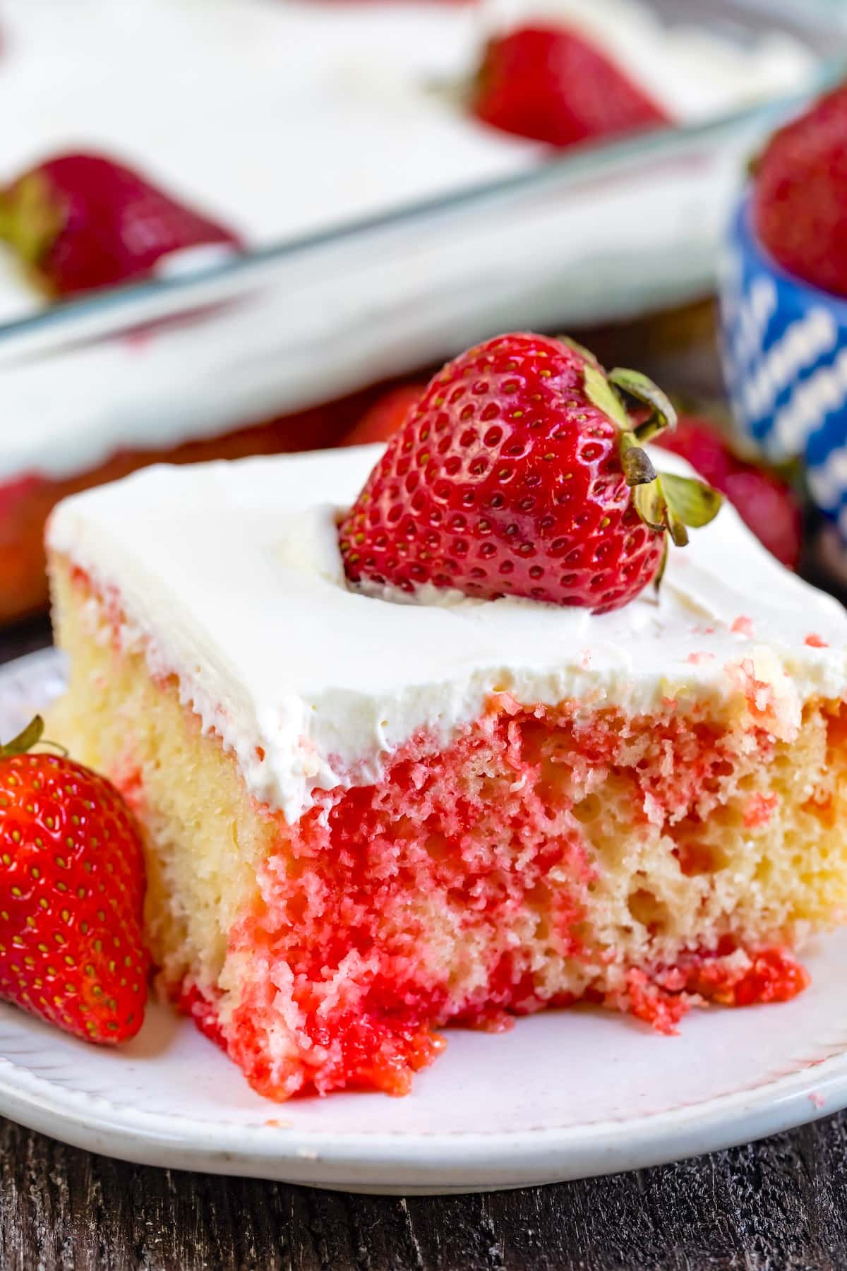 A slice of strawberry poke cake with white frosting and a whole strawberry on top, sitting on a white plate. The cake has visible red strawberry gelatin throughout and fresh strawberries beside it.