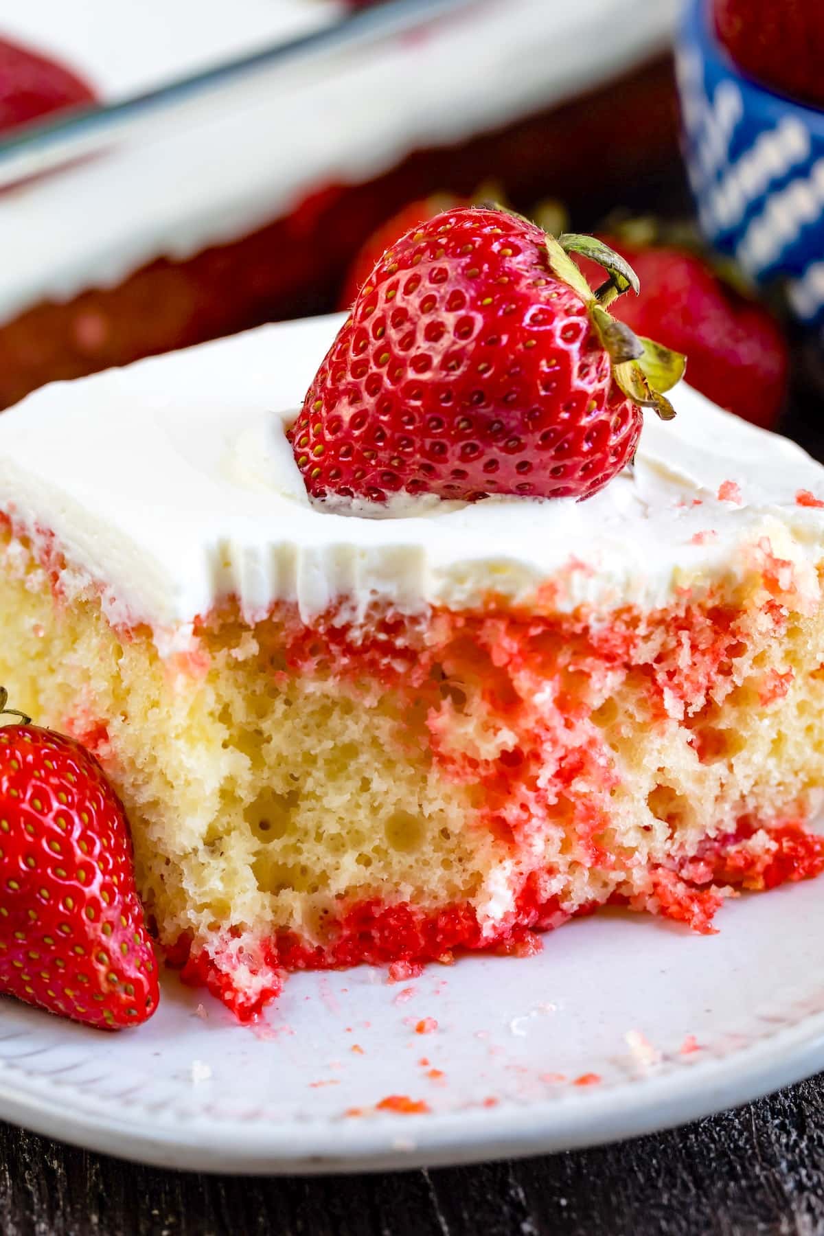 A close-up of a slice of strawberry poke cake with white frosting, topped with a fresh strawberry. The cake is moist with red strawberry sauce visible inside and around the slice, and another whole strawberry is on the plate.
