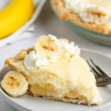 A slice of banana cream pie topped with whipped cream and banana slices is on a plate with a fork. In the background, there is a whole banana and the remainder of the pie.