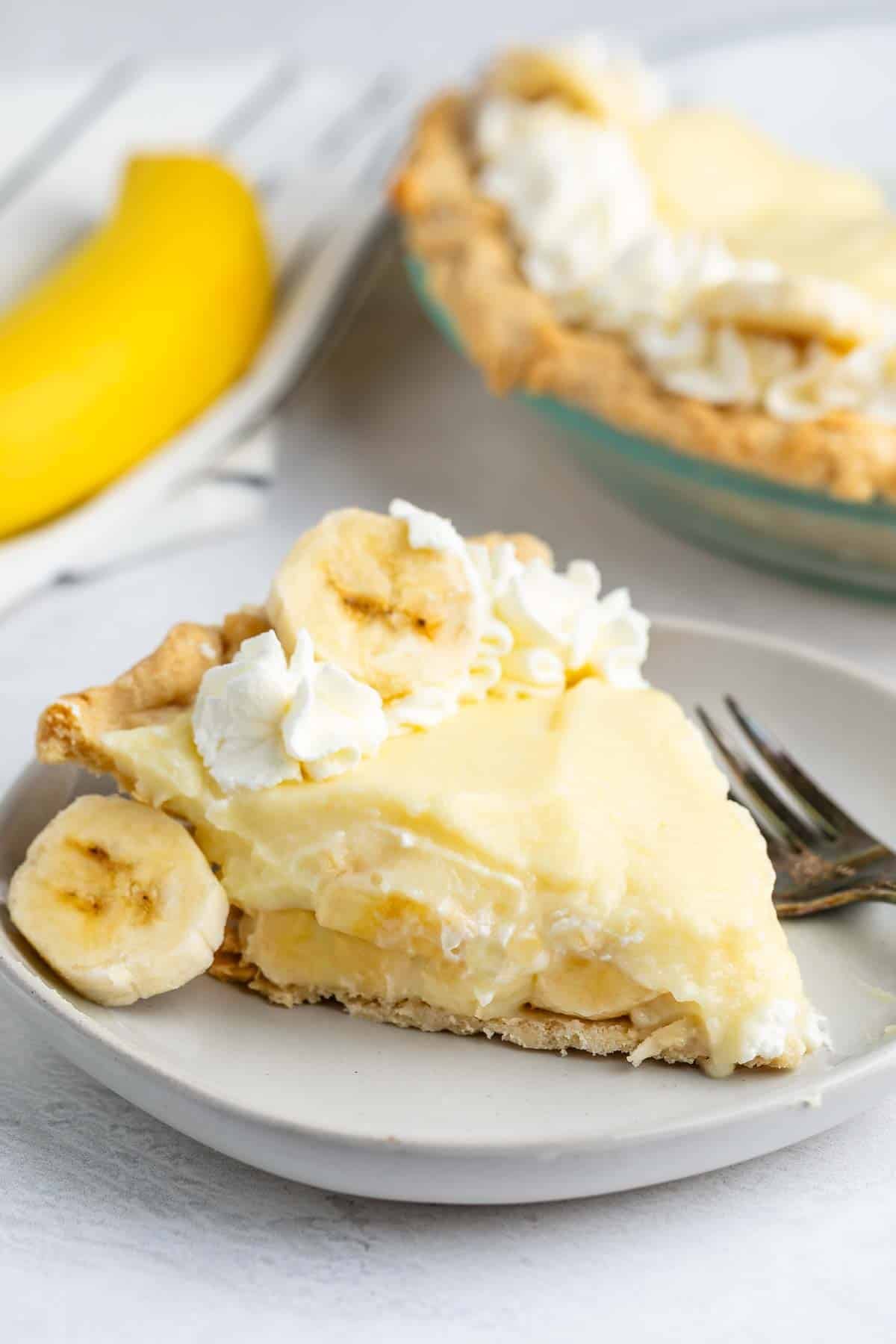 A slice of banana cream pie topped with whipped cream and banana slices is on a plate with a fork. In the background, there is a whole banana and the remainder of the pie.