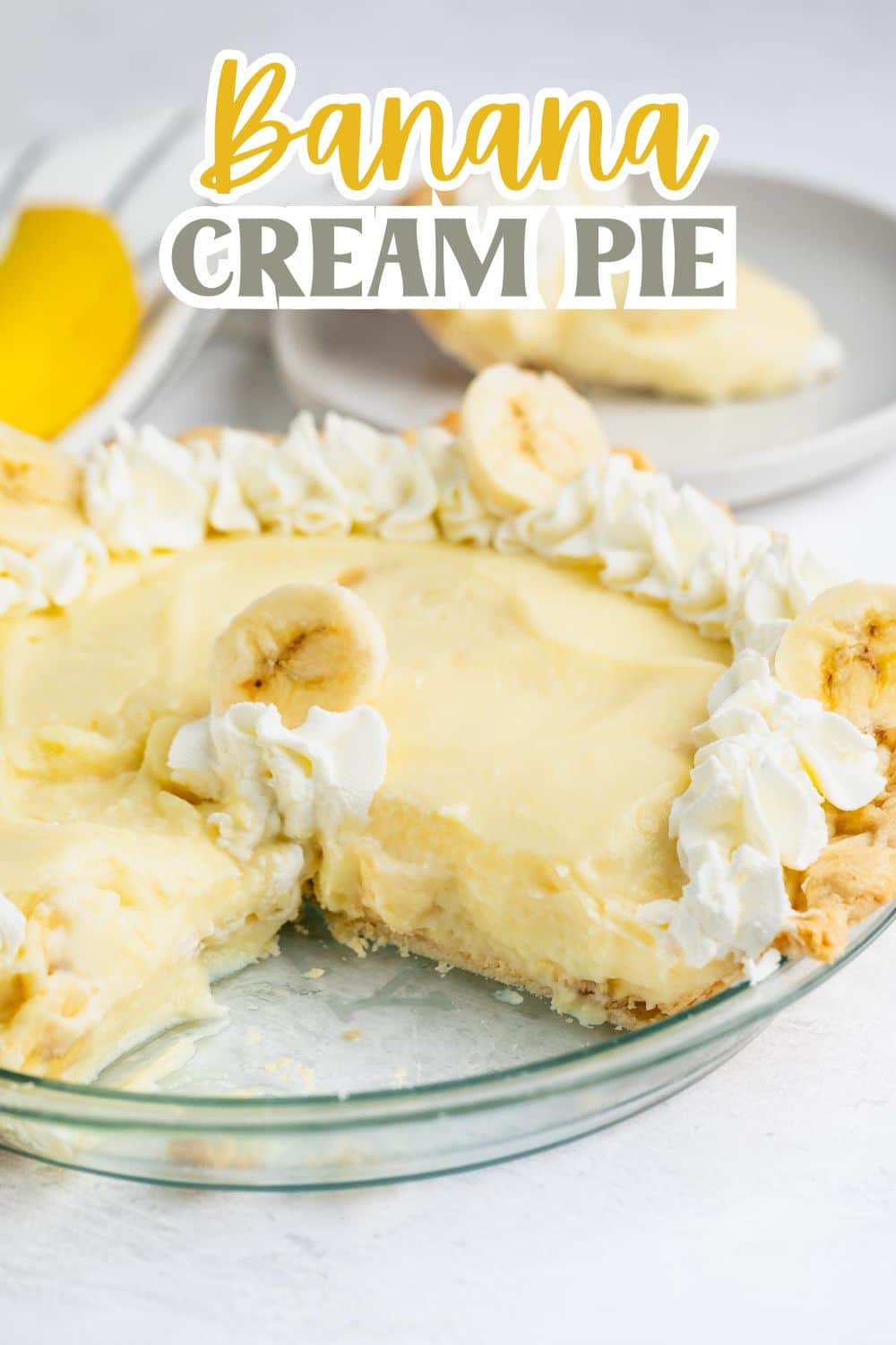 A banana cream pie with a slice missing, topped with whipped cream and banana slices, sits in a glass pie dish. A plate with a serving of the pie and a banana are in the background. Text reads Banana Cream Pie.