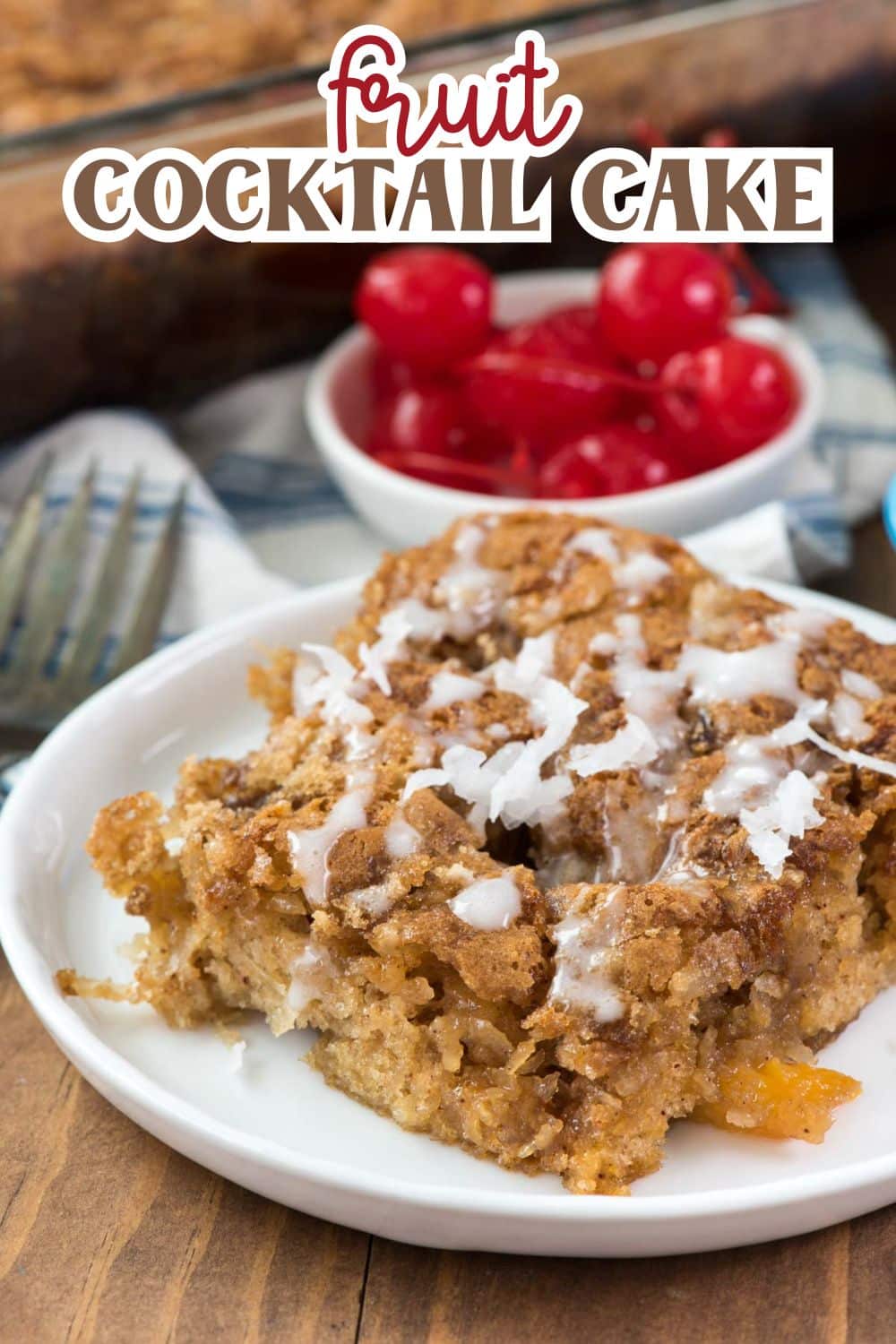 A slice of fruit cocktail cake with white icing sits on a white plate. In the background, a bowl of red maraschino cherries and a baking dish are visible. The words fruit COCKTAIL CAKE are at the top of the image.