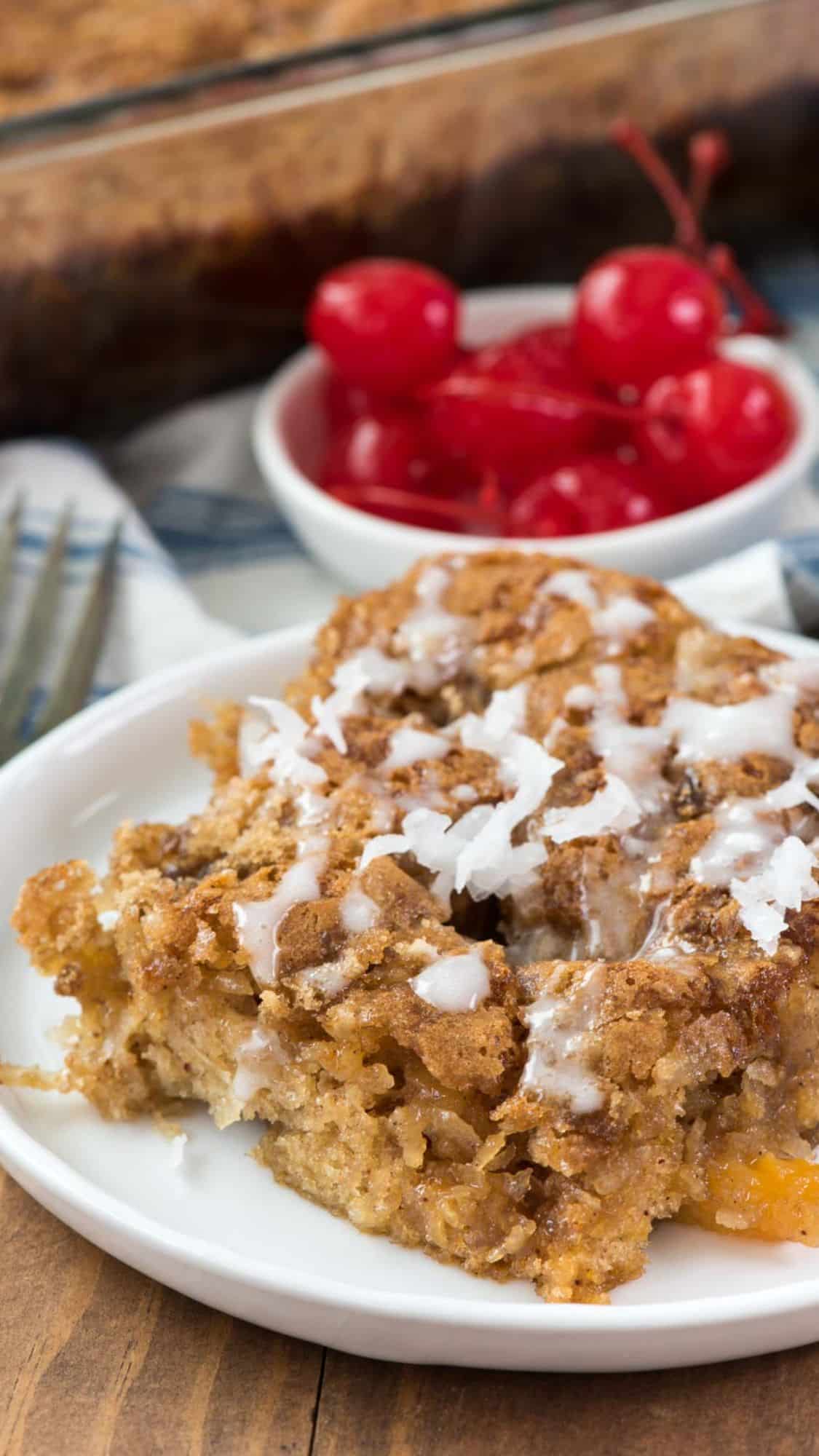 A slice of crumbly coffee cake with white icing on a white plate, served in front of a small bowl of bright red cherries and a baking dish in the background.